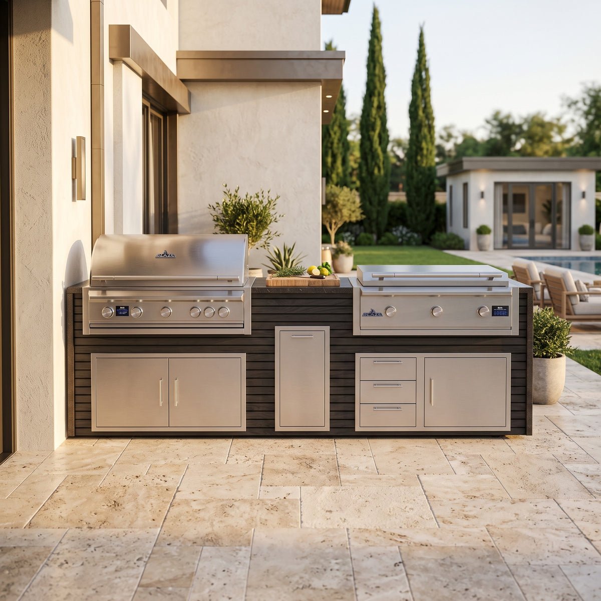Cindered Ash grill and griddle island with Charred Onyx granite top on a travertine patio against the house with cypress trees and a pool house beyond