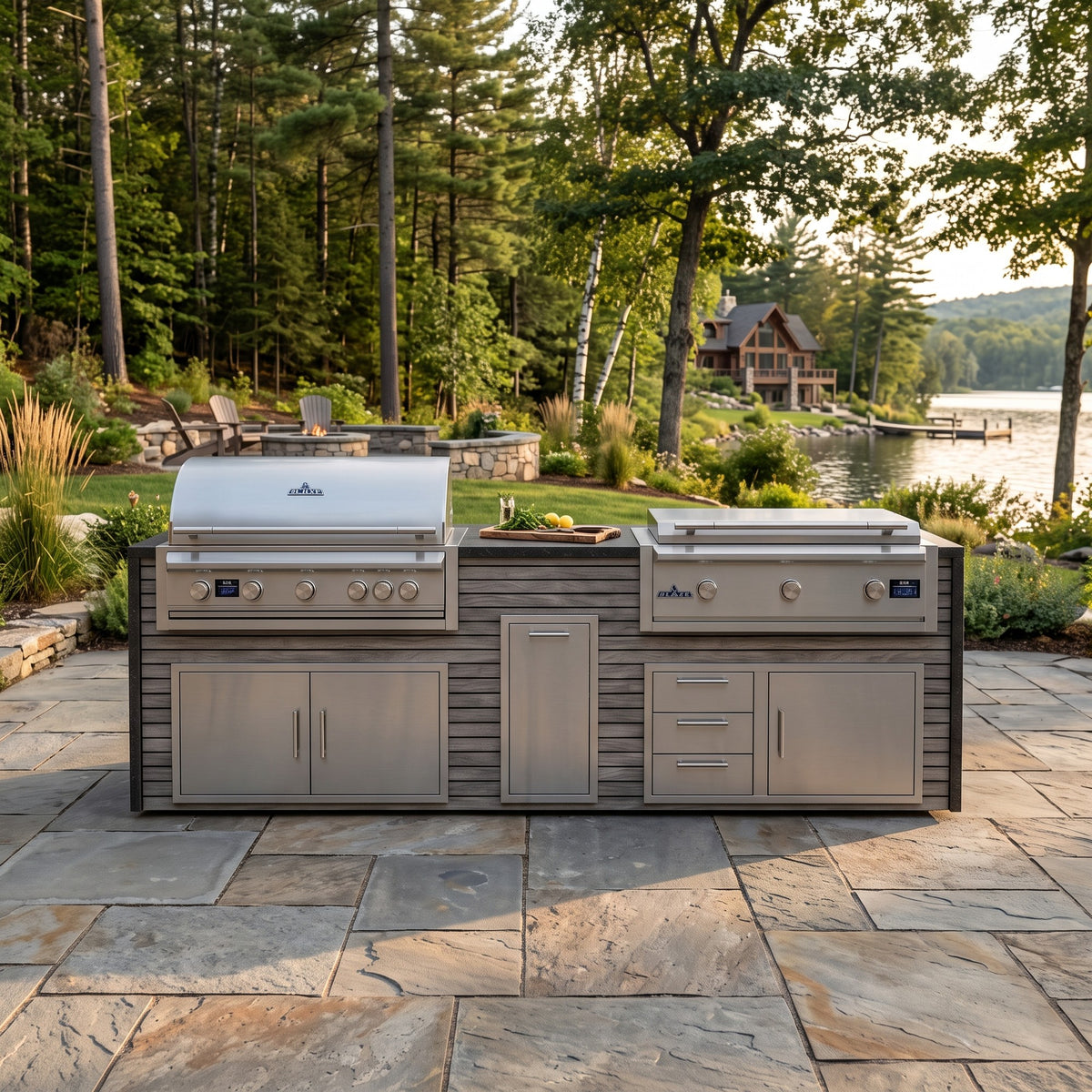 Drifted Cypress grill and griddle island with Charred Onyx granite top on a flagstone patio by a lake with a wooded shoreline and lake house