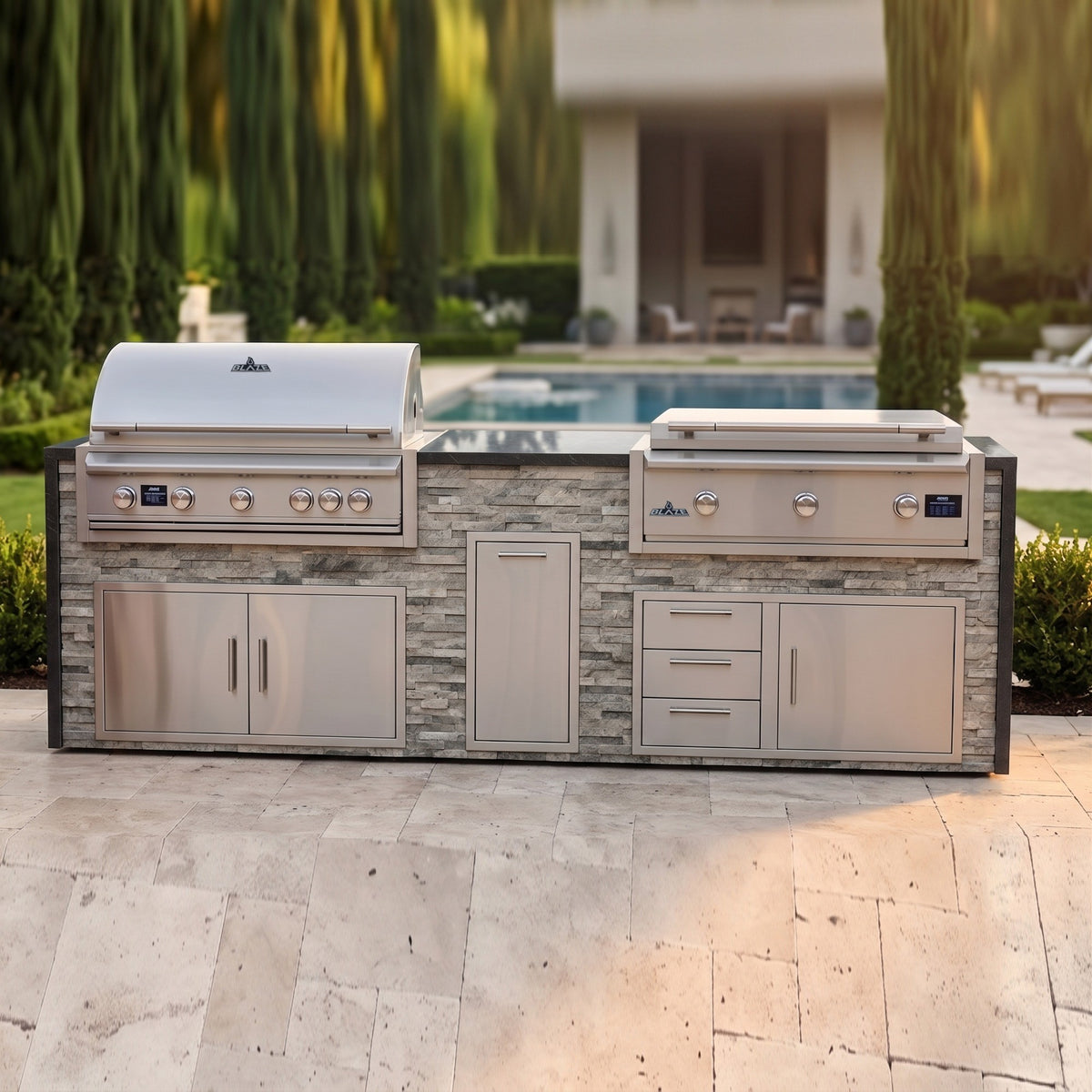 Tuscan Hearth grill and griddle island with Charred Onyx granite top on a travertine patio with the pool and pool house behind tall cypress trees