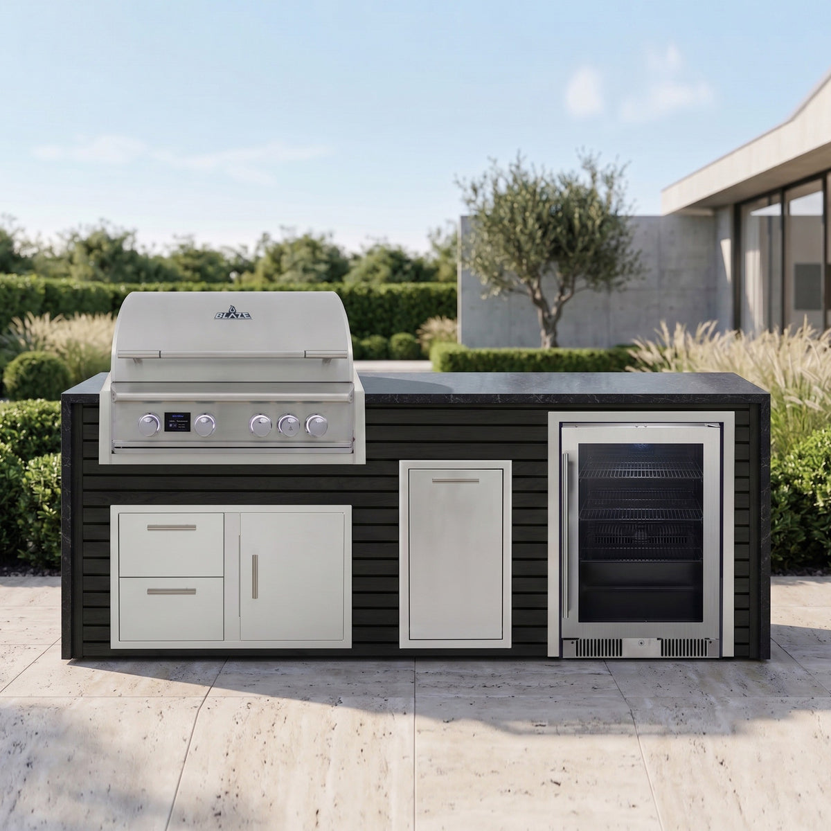 Cindered Ash outdoor kitchen with beverage cooler and Charred Onyx granite top on a travertine patio with ornamental grasses, an olive tree and a modern concrete home