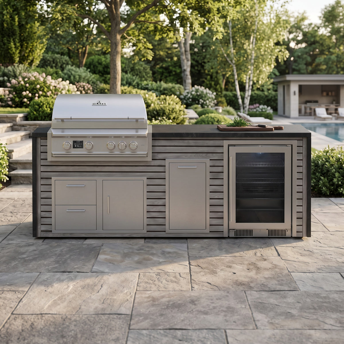 Drifted Cypress outdoor kitchen with beverage cooler and Charred Onyx granite top on a flagstone patio surrounded by garden beds with a pool house beyond