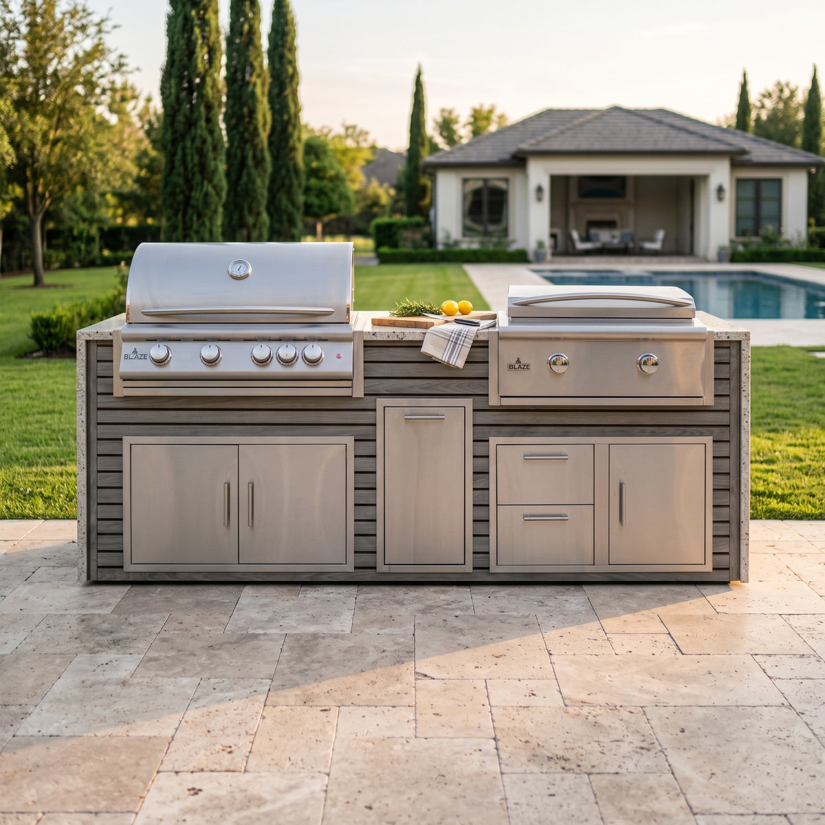 Drifted Cypress grill and griddle island with Frosted Ember granite top on a travertine patio facing the lawn, pool and pool house