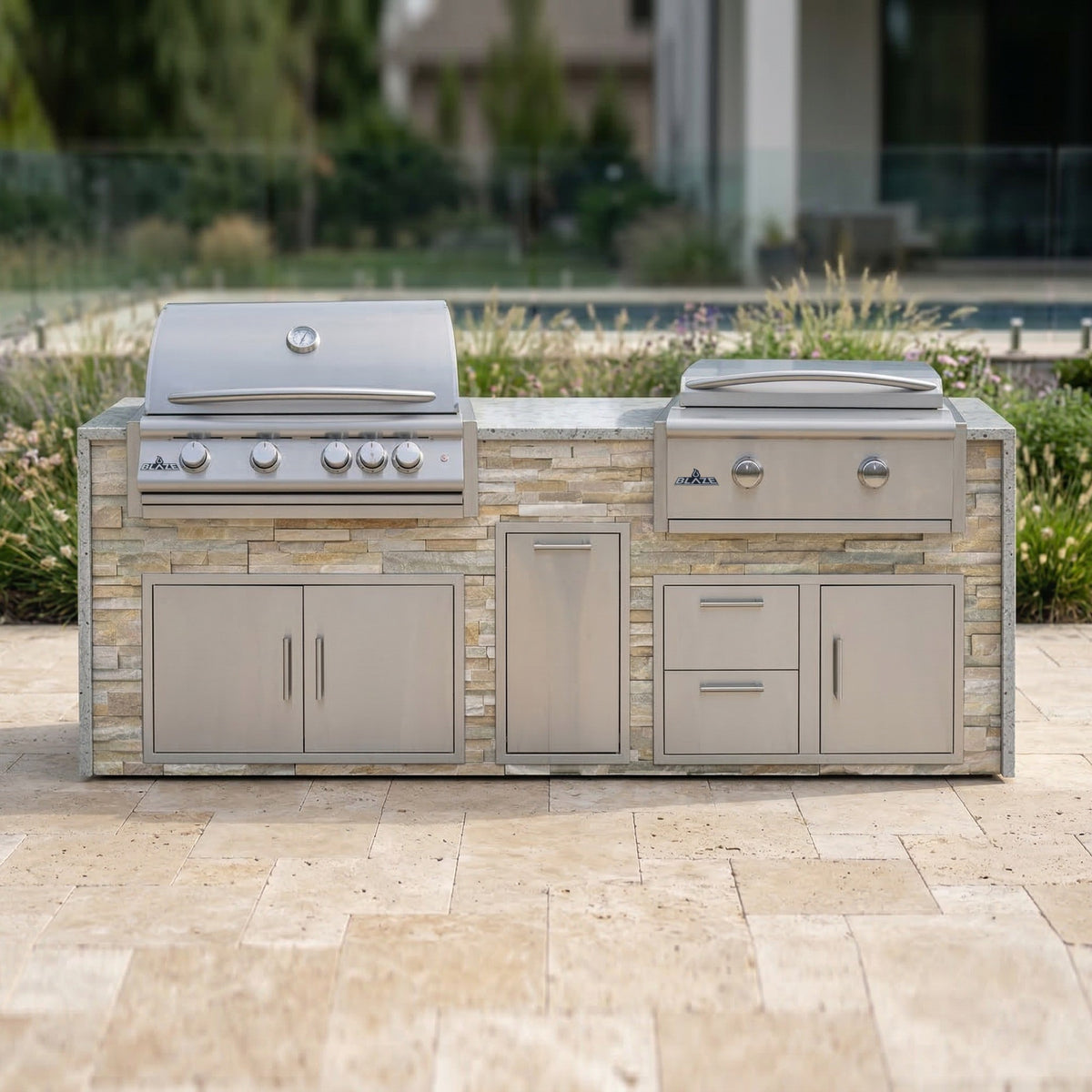 Sunkissed Dune grill and griddle island with Frosted Ember granite top on a travertine patio with ornamental grasses and a pool behind glass fencing