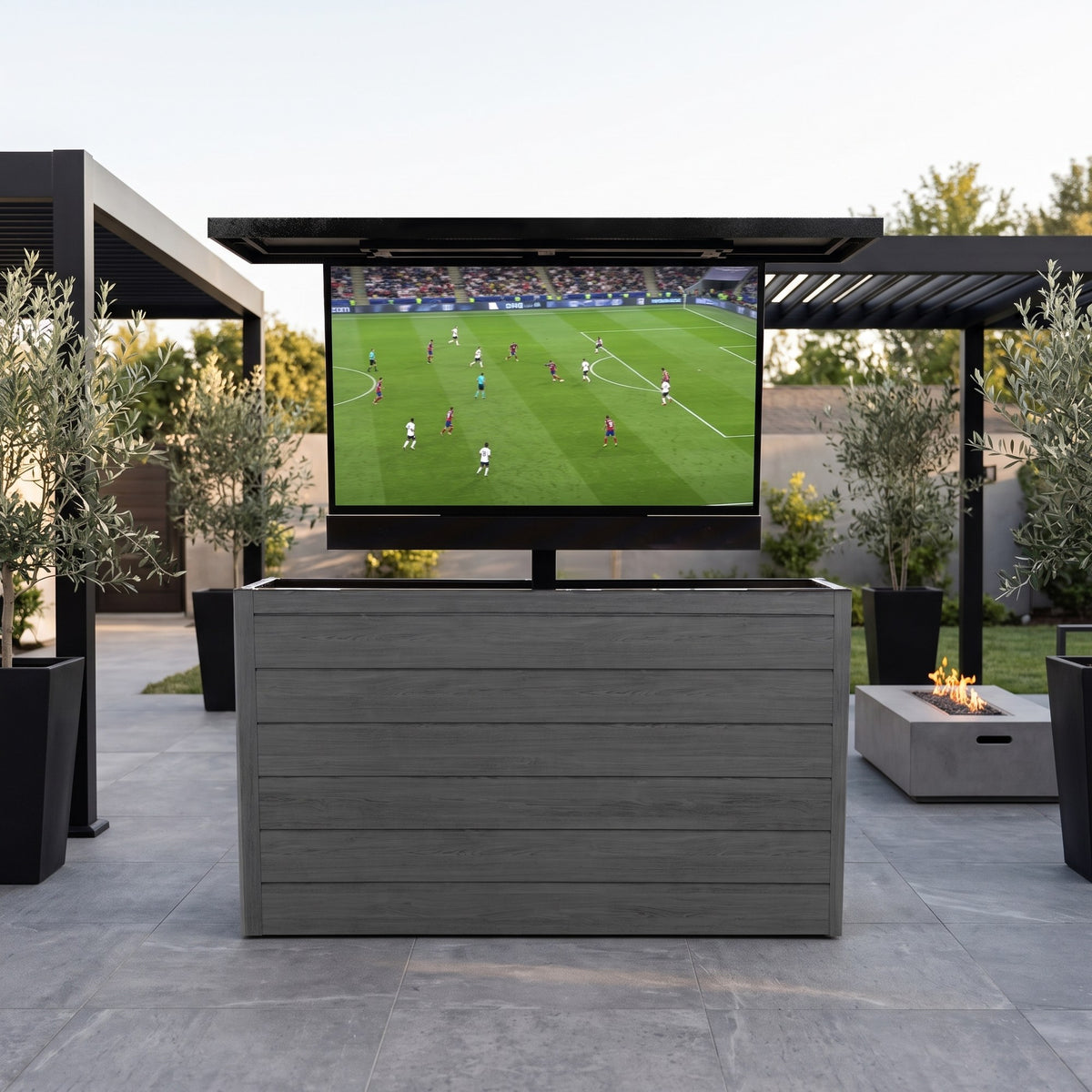 Cindered Ash 6 inch ArtisanPlank outdoor TV lift cabinet with the TV raised on a modern patio with olive trees and a concrete fire table