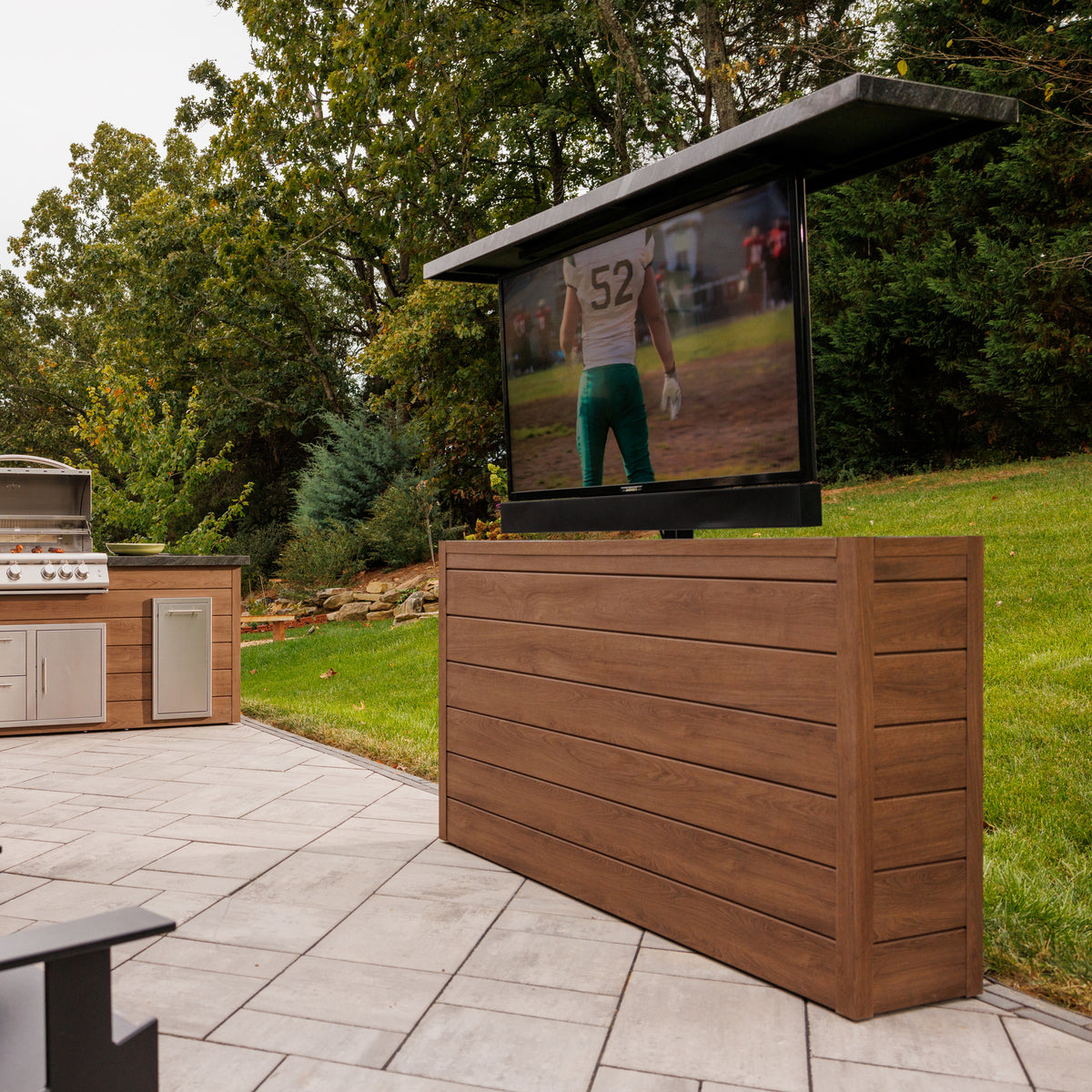 Umbered Oak 6 inch ArtisanPlank outdoor TV lift cabinet with the TV raised on a paver patio beside an outdoor kitchen