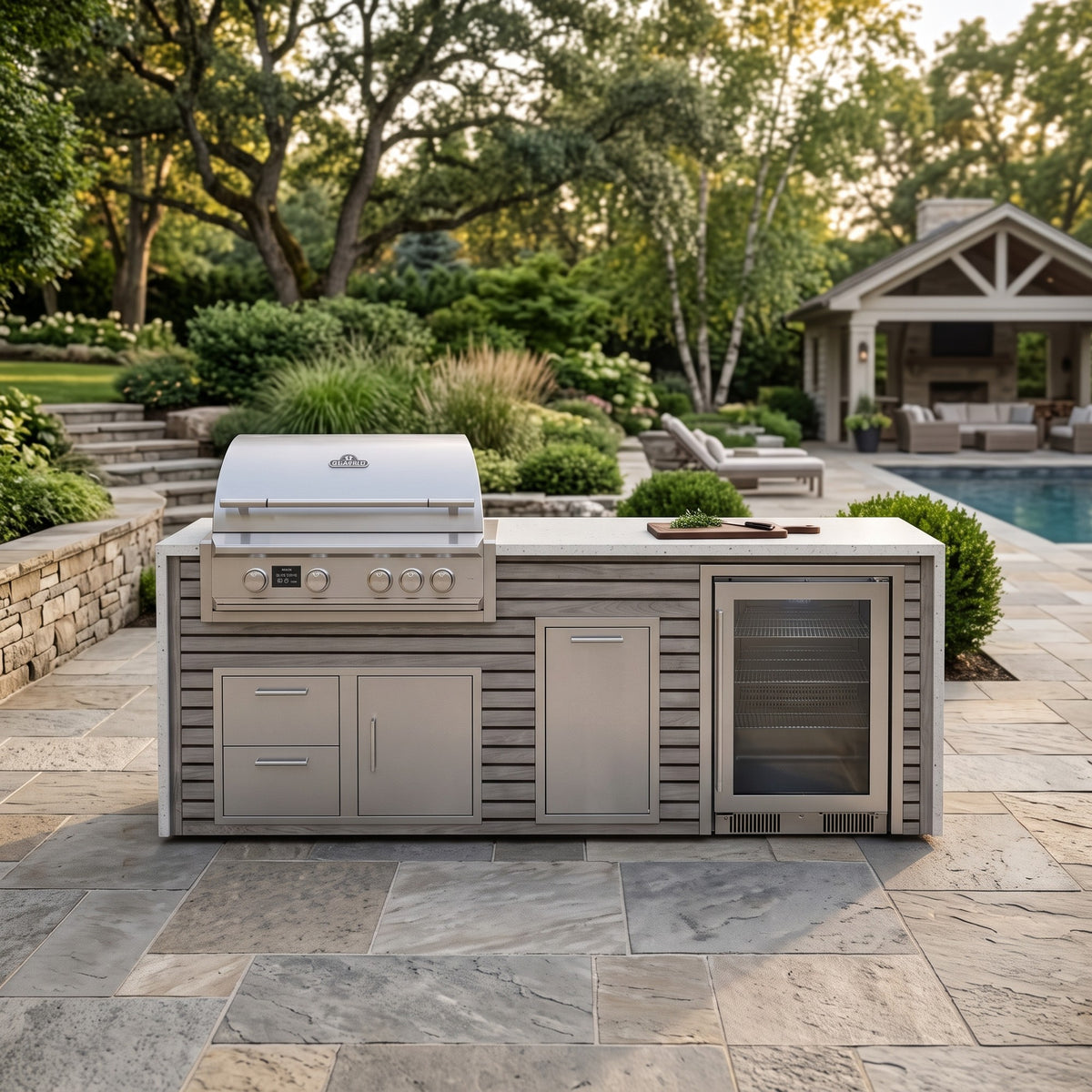 Drifted Cypress outdoor kitchen with beverage cooler and Frosted Ember granite top on a flagstone patio facing the pool and a covered pavilion under shade trees