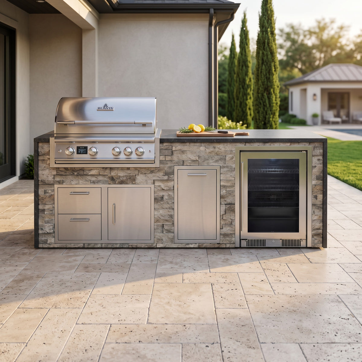 Tuscan Hearth outdoor kitchen with beverage cooler and Charred Onyx granite top on a travertine patio beside the house with cypress trees and a pool house beyond