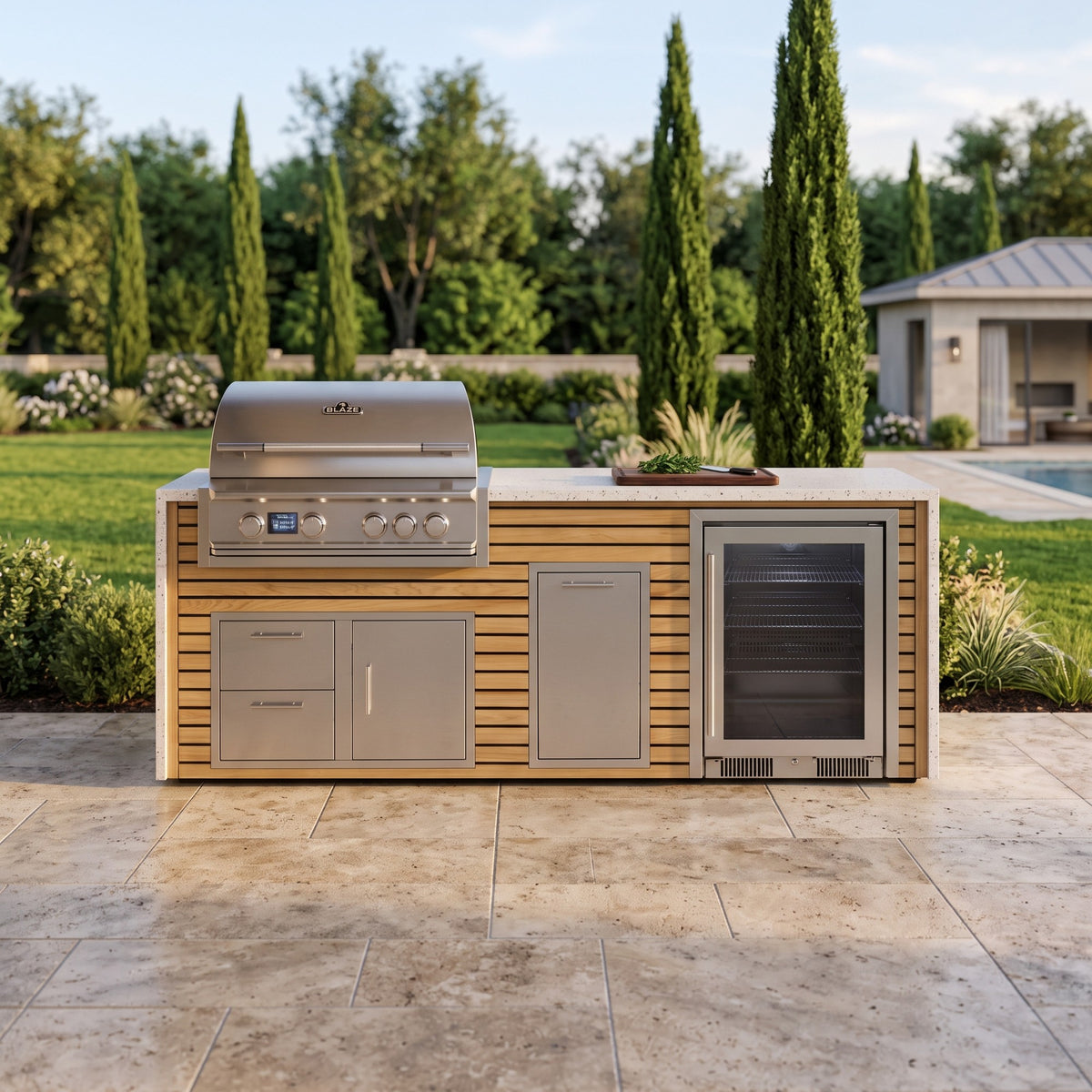 Warmed Acacia outdoor kitchen with beverage cooler and Frosted Ember granite top on a travertine patio edged with lawn and flower beds, pool house behind